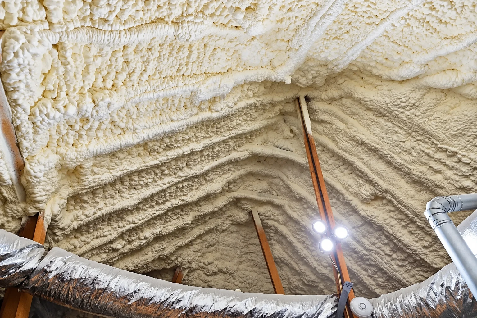 Thick, uneven white spray foam coats the sloped attic roof around exposed wooden beams and ductwork.
