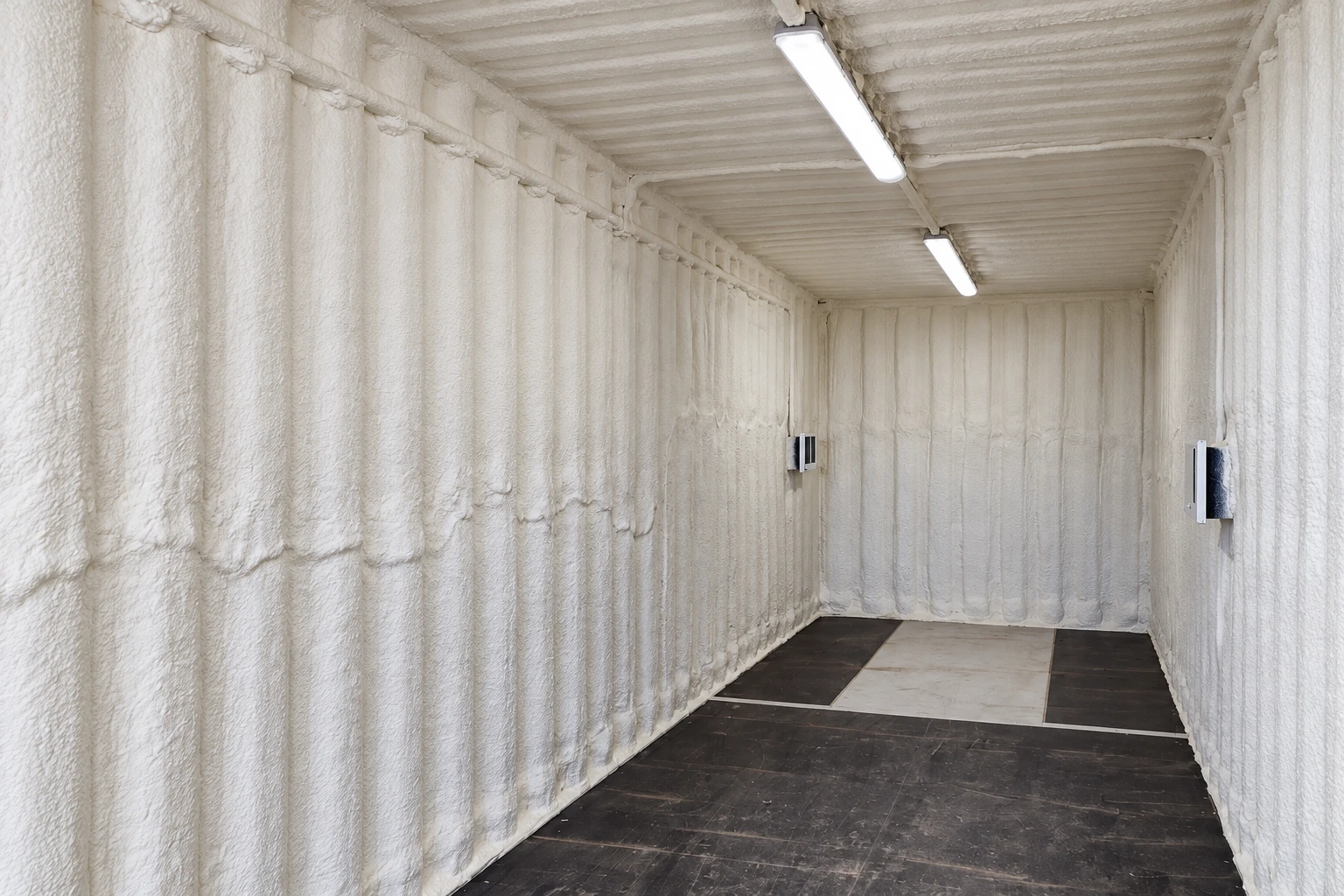 Commercial spray foam insulation covers the walls and ceiling of a long, empty room lit by overhead fixtures.