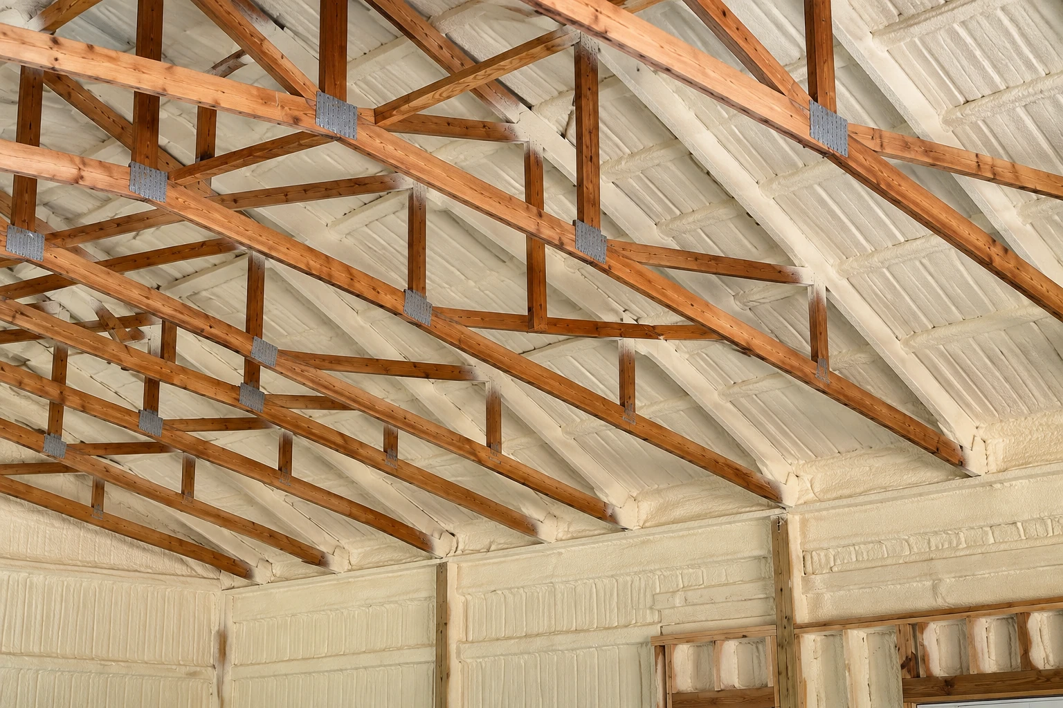 Spray foam insulation coats the roof and walls beneath exposed wooden trusses in a large building.