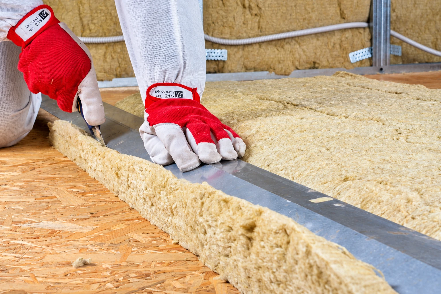 A gloved worker cuts a tan insulation batt to fit between metal framing on a plywood floor.