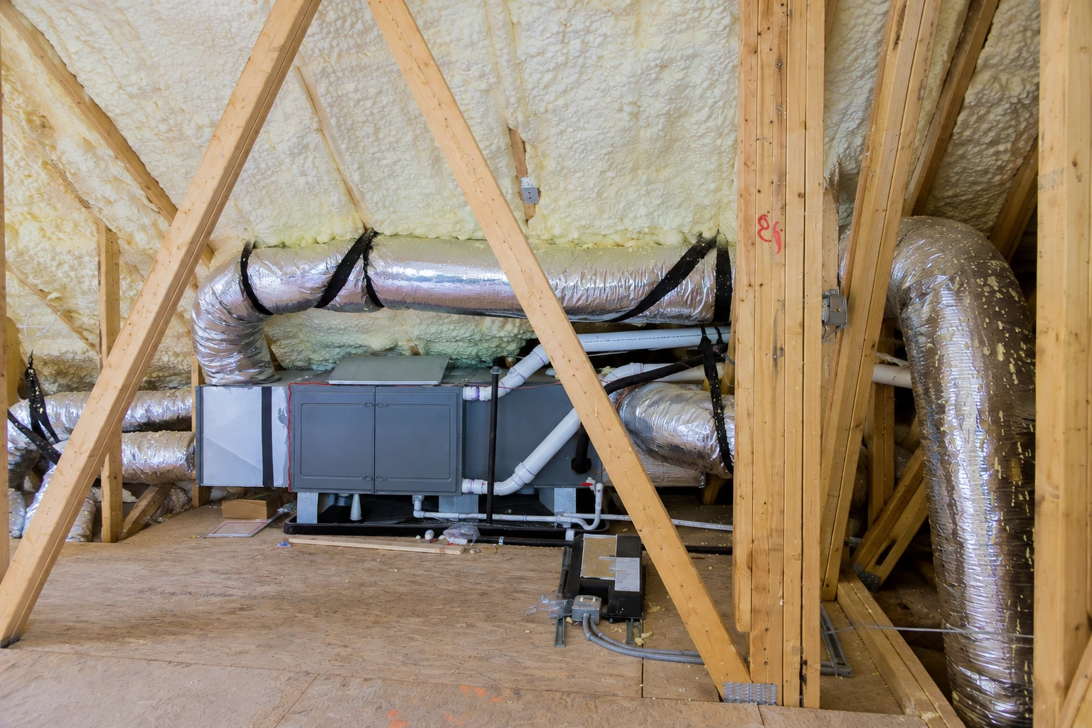 Open-cell foam insulation coats an attic roof around exposed wooden trusses, ductwork, and an HVAC unit.