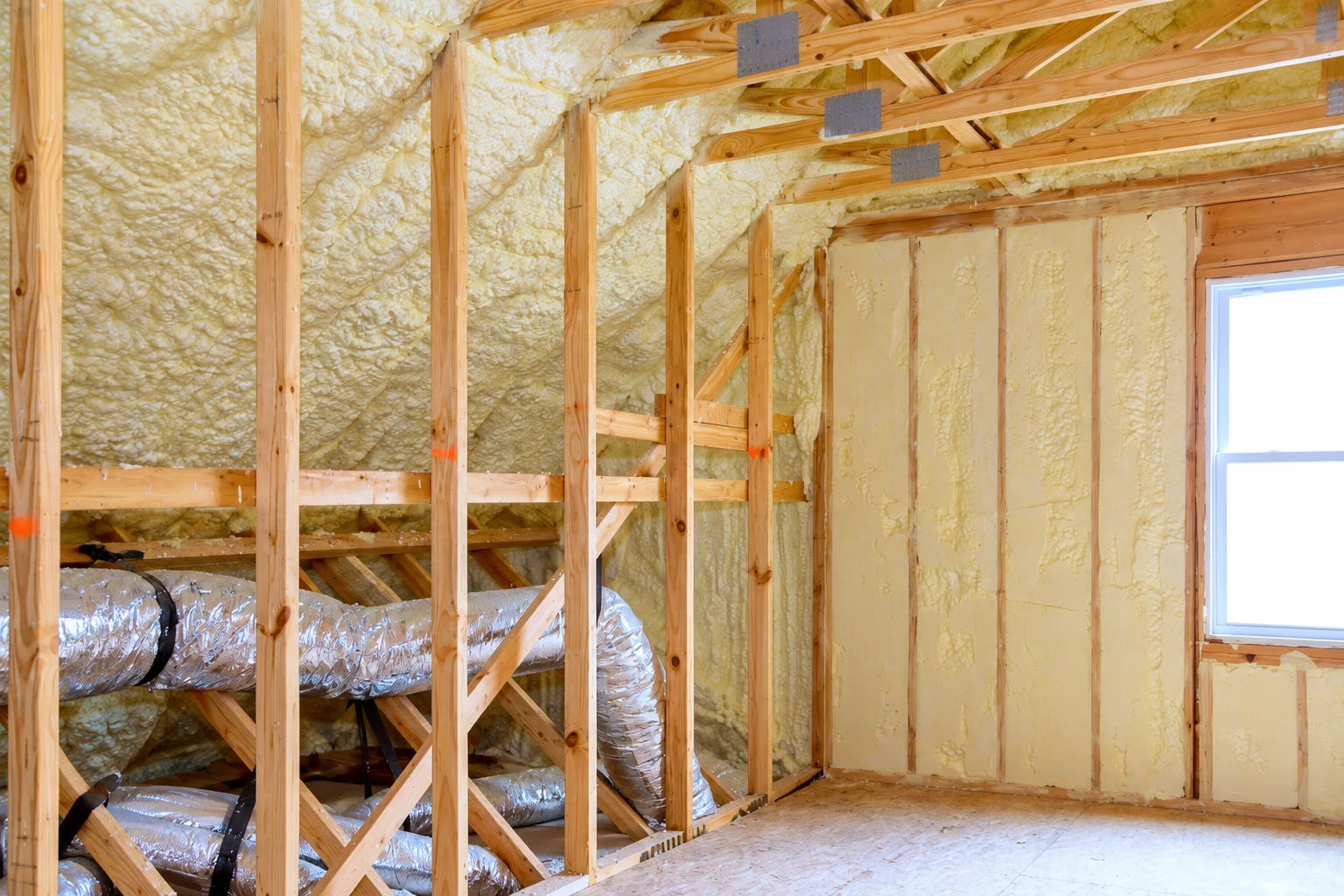 Foam insulation fills the sloped roof and wall framing in an unfinished room, with exposed ducts and a window.