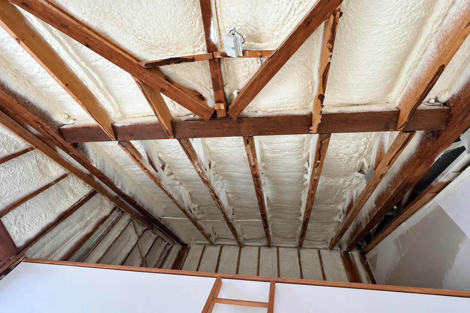 Spray foam insulation covers the roof underside between exposed brown wooden rafters in an attic.