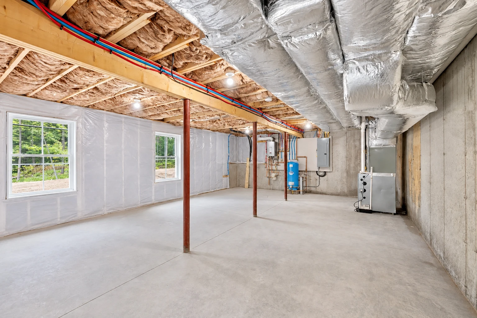 Vapor barrier lines a basement wall above a concrete floor, beneath exposed joists and large silver ducts.