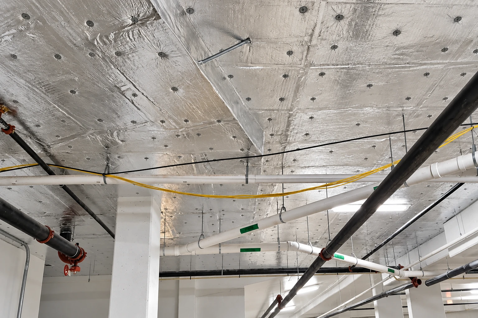 Reflective vapor barrier panels cover a commercial ceiling around pipes, cables, and support columns.