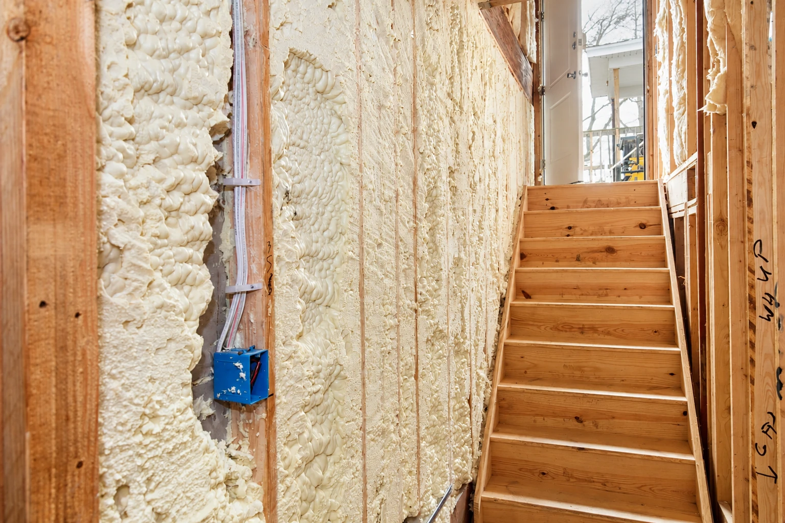 Spray foam coats exposed wall framing beside a wooden staircase in an unfinished interior.